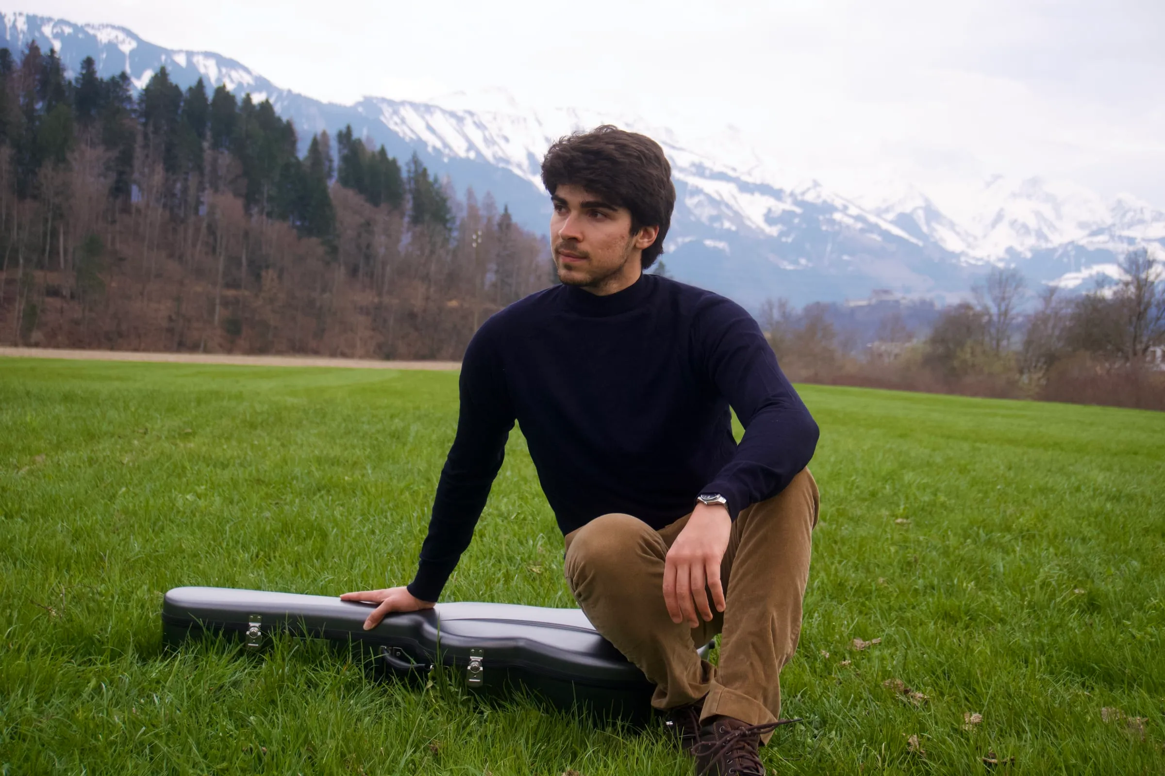 Miguel Ferreira sitting on a meadow beside his guitar case, snow-covered mountains behind him.