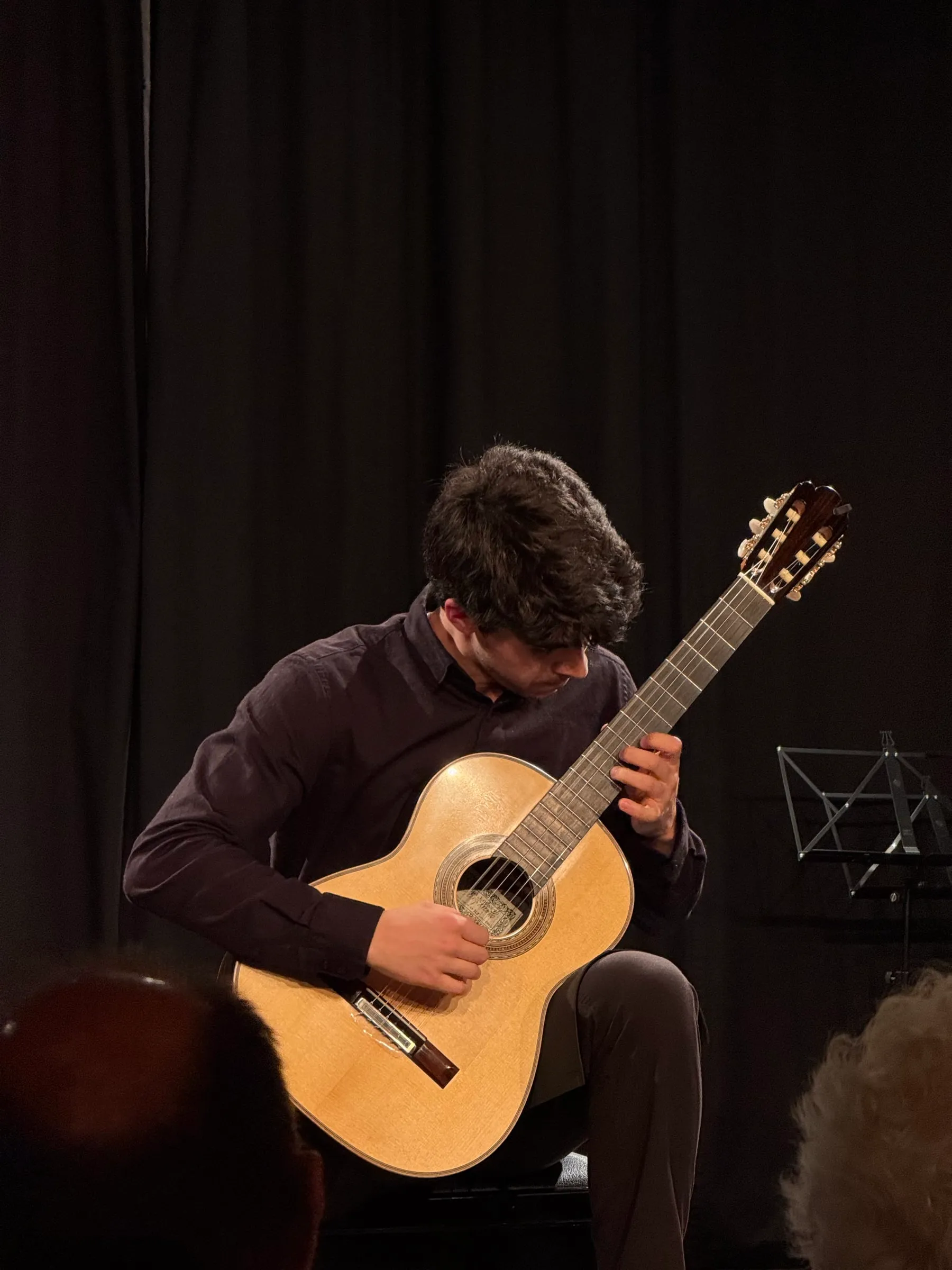 Miguel Ferreira performing on the dark stage of the Teatro Comico in Sion, head bowed over his classical guitar under a warm spotlight, audience silhouettes in the foreground.